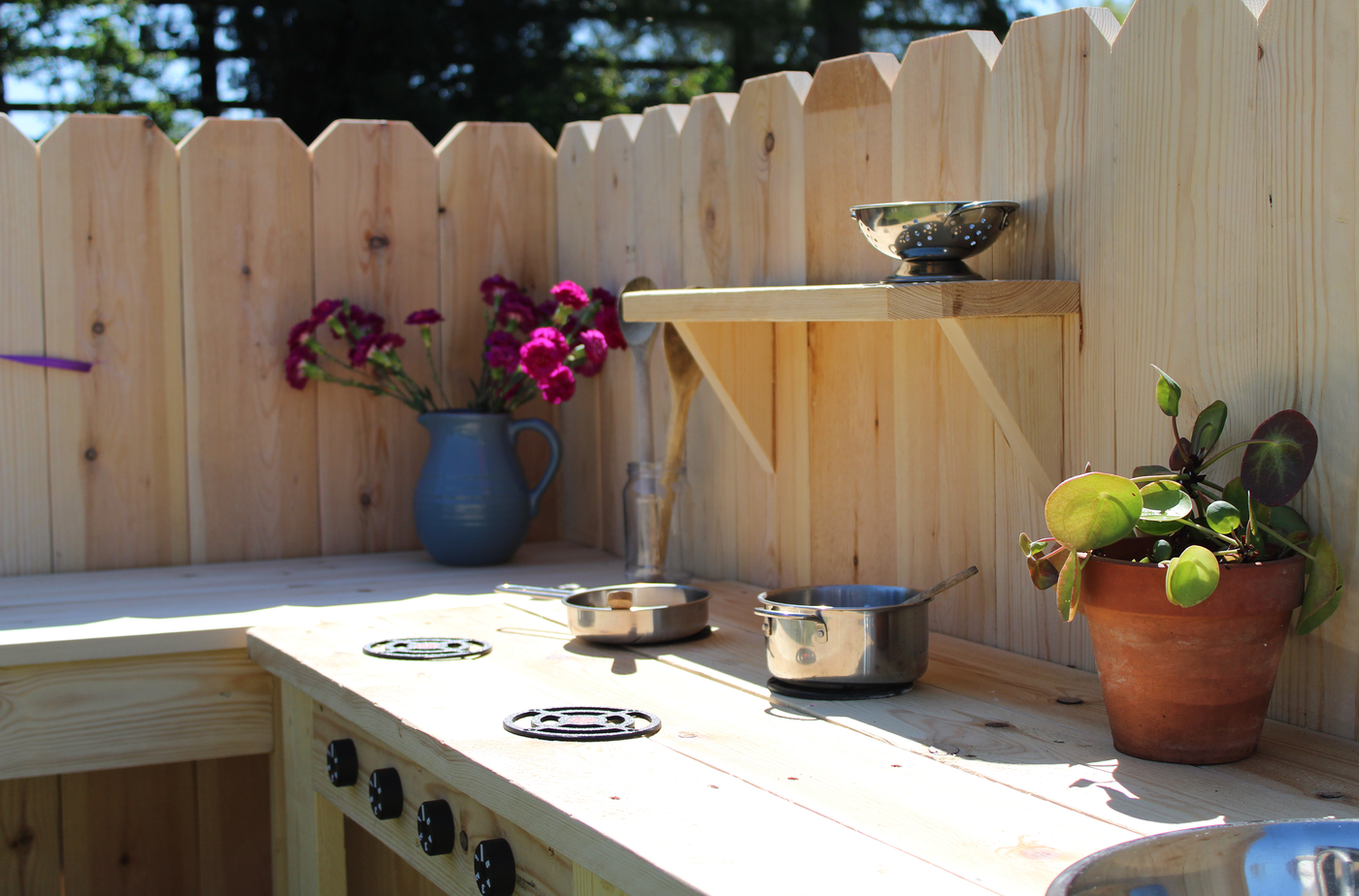 Outdoor wooden kitchen setup with pots, pans, and decorative items against a wooden fence.