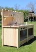 Wooden play kitchen set outdoors on a grassy area with trees and a playground in the background.