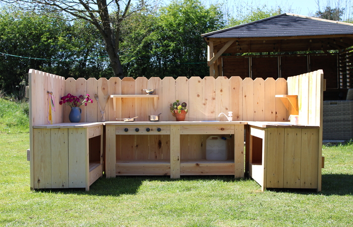 Wooden outdoor kitchen setup with shelves and a sink in a grassy area.