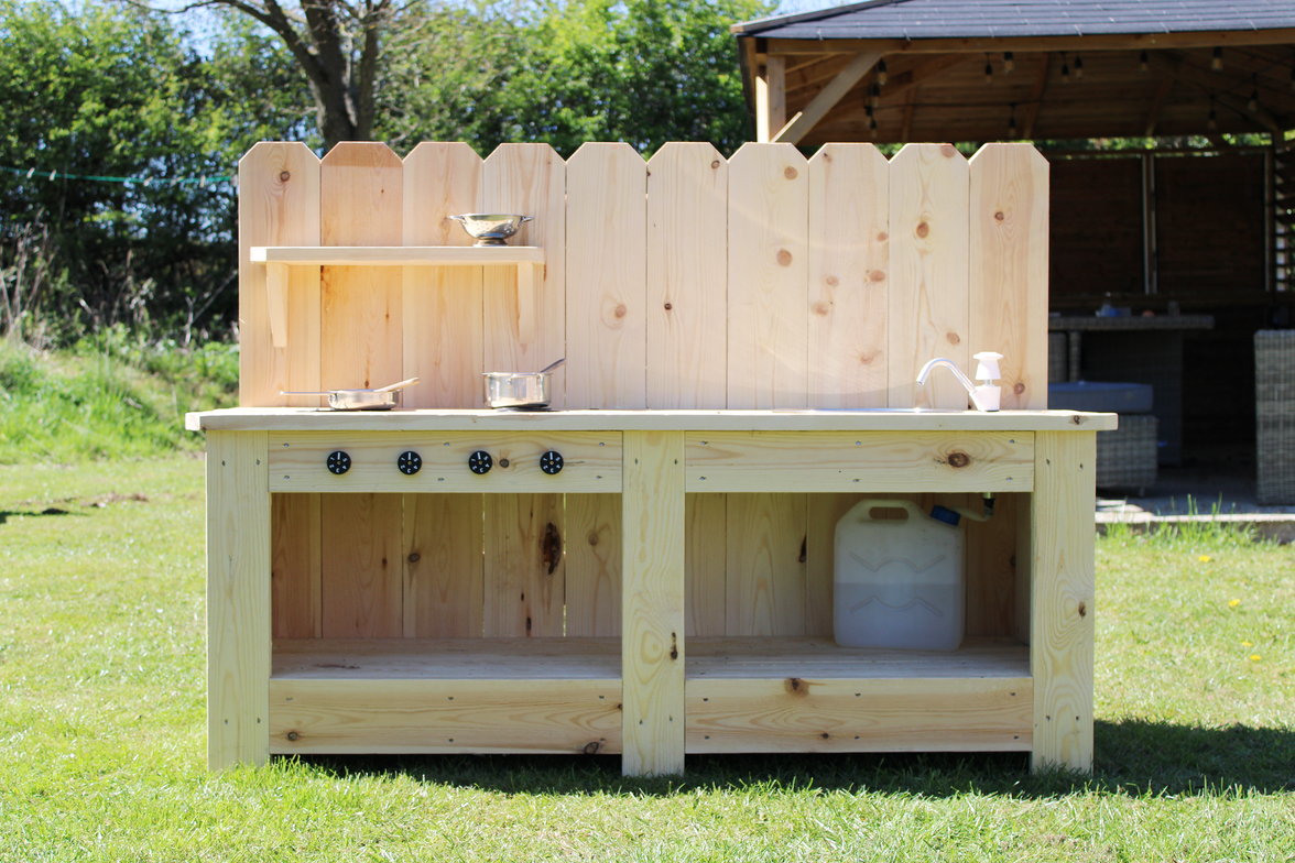 Wooden outdoor kitchen with shelves and cooking utensils on a grassy area.