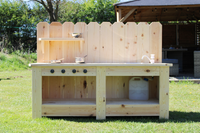 Wooden outdoor kitchen with shelves and cooking utensils on a grassy area.