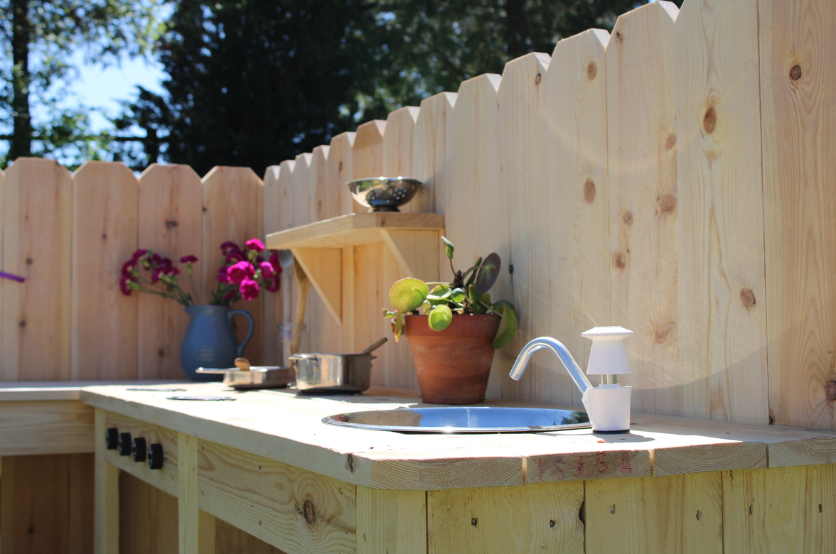 Outdoor wooden kitchen setup with sink, pots, and flowers against a wooden fence.
