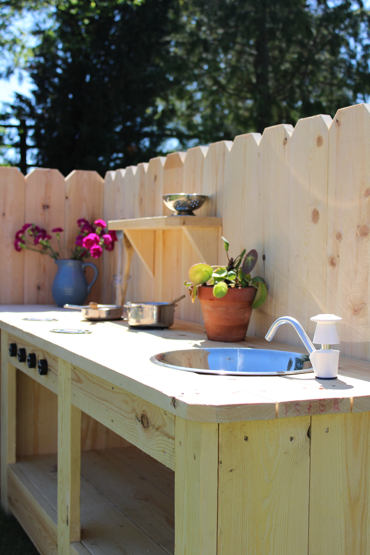 Outdoor kitchen setup with wooden counter, sink, and potted plants against a wooden fence.