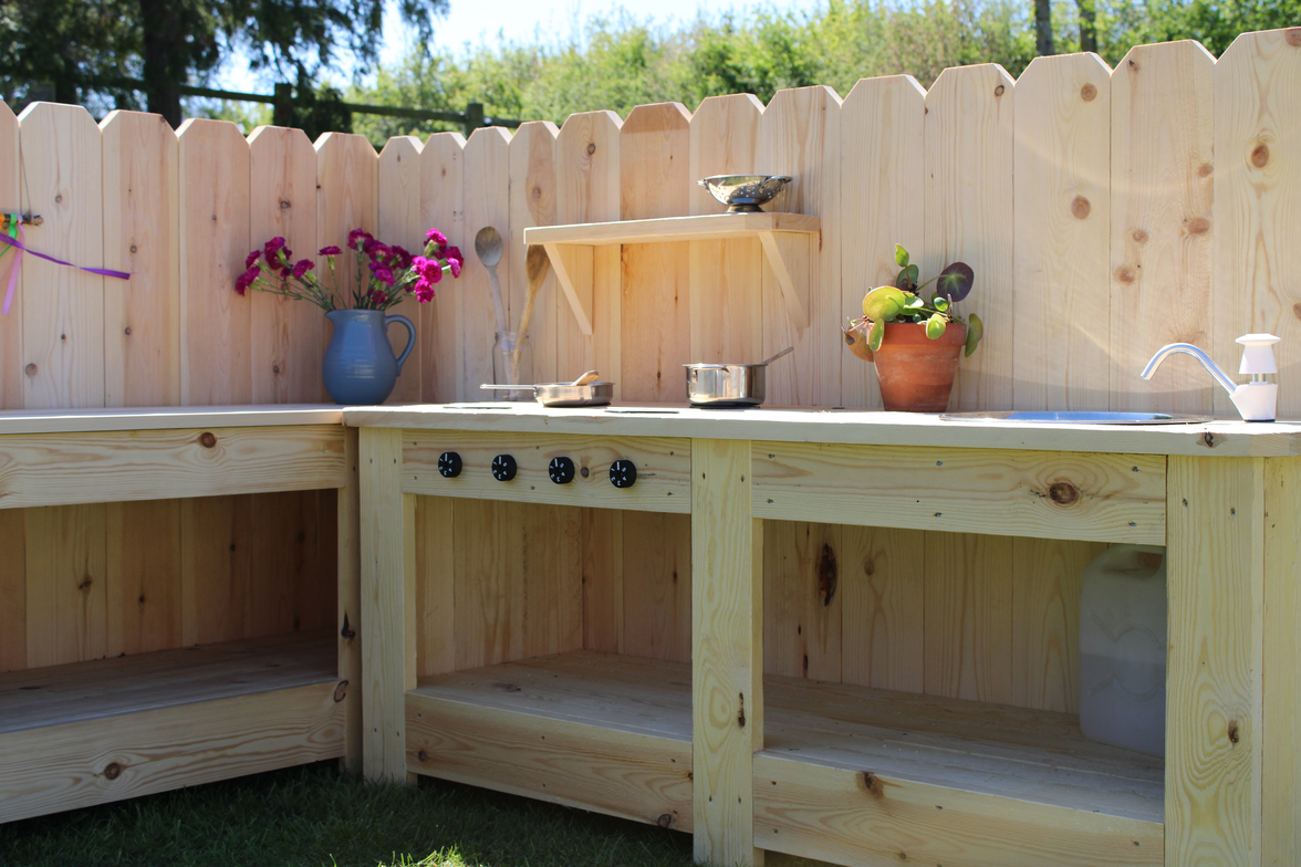 Wooden outdoor play kitchen with shelves and decorative elements against a wooden fence.