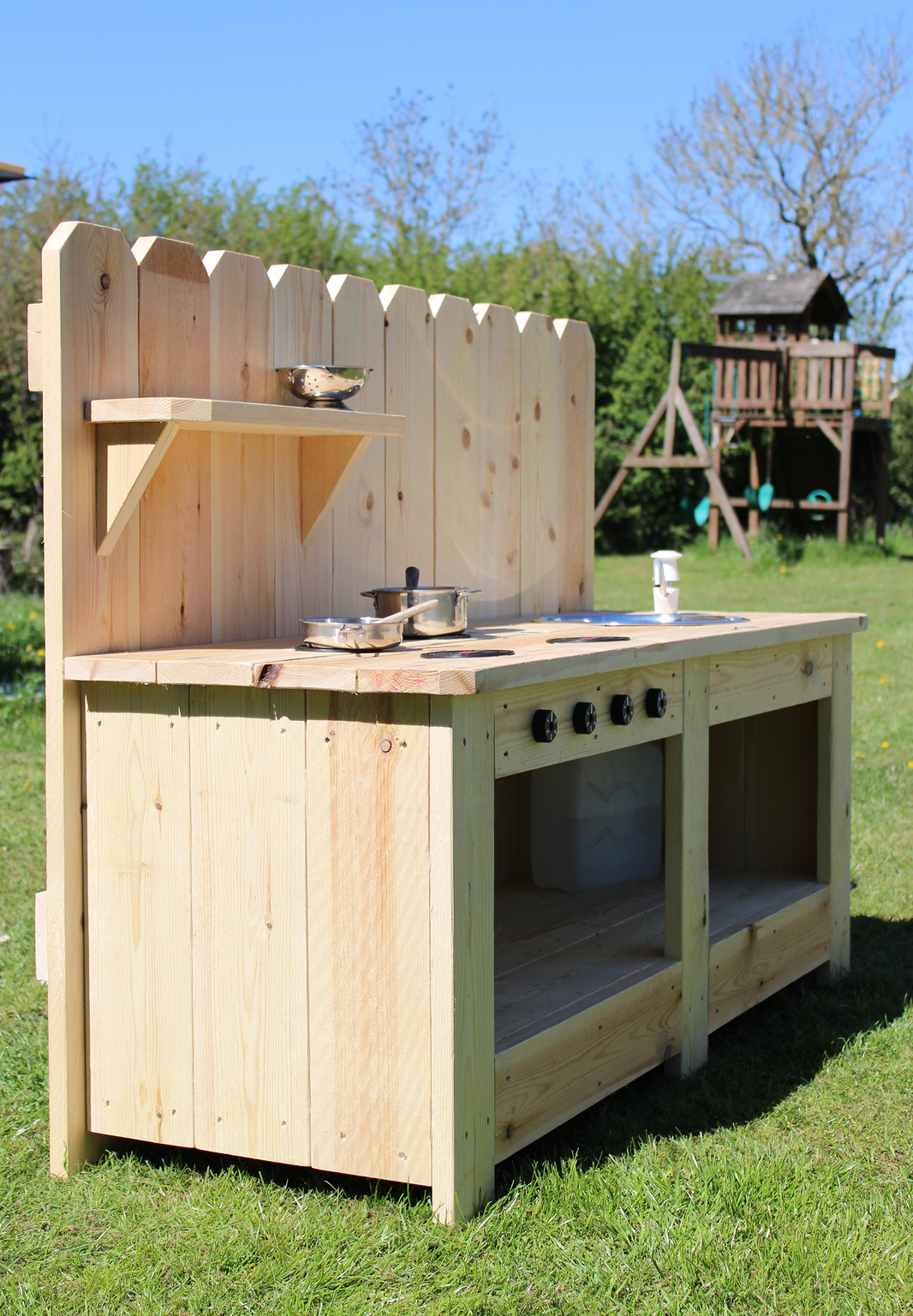 Wooden play kitchen set outdoors on a grassy area with trees and a playground in the background.