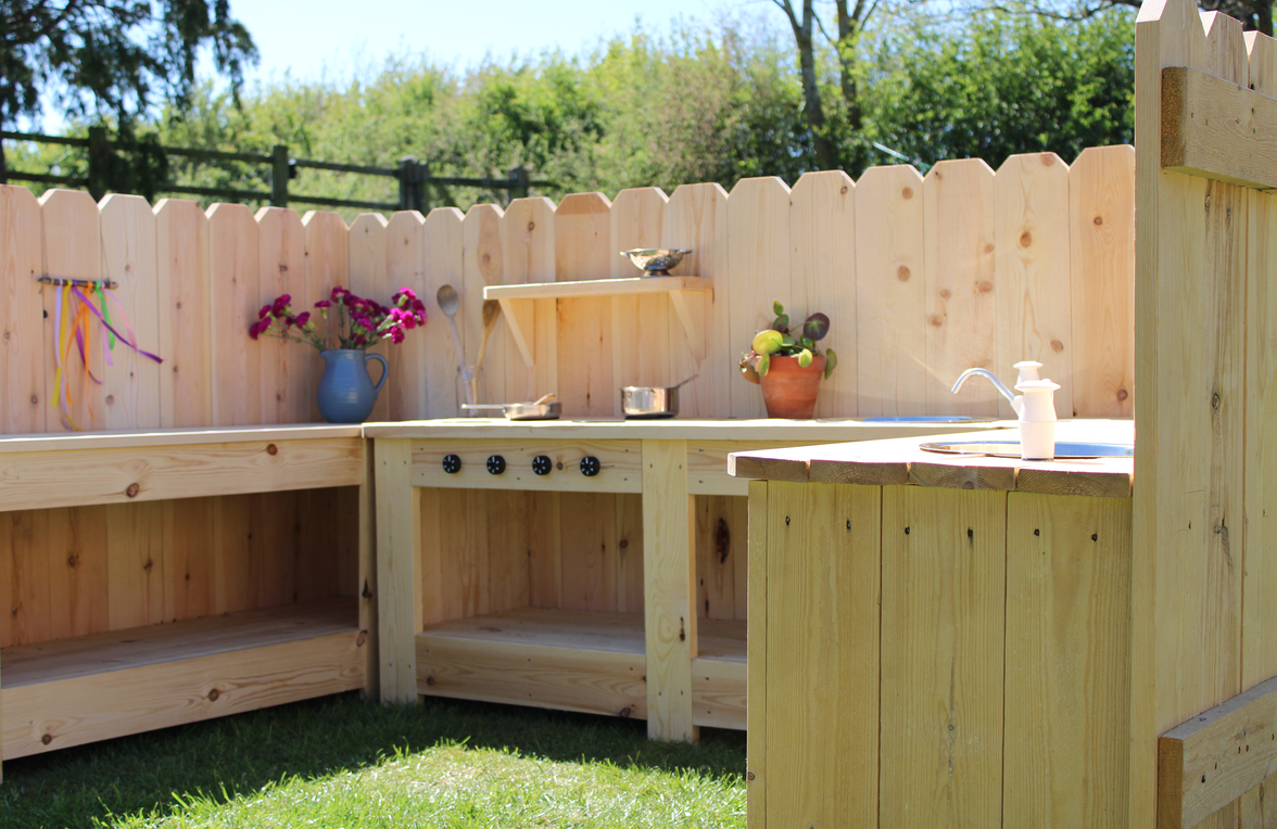 Wooden outdoor kitchen with sinks and shelves against a wooden fence.