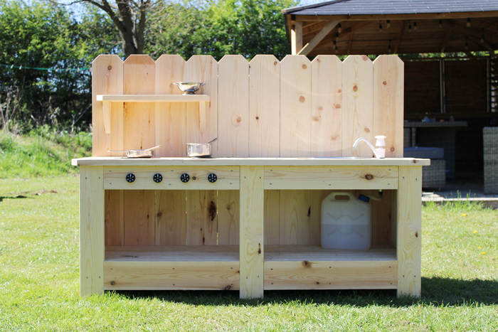 Wooden outdoor kitchen with shelves and cooking utensils on a grassy area.